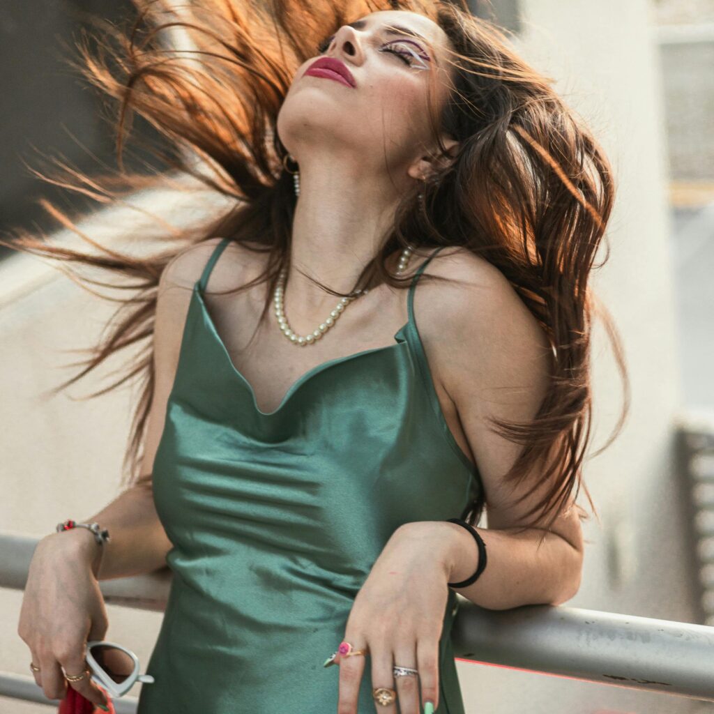 A stylish woman in a green dress poses outdoors, her hair blowing in the wind, exuding confidence and grace.