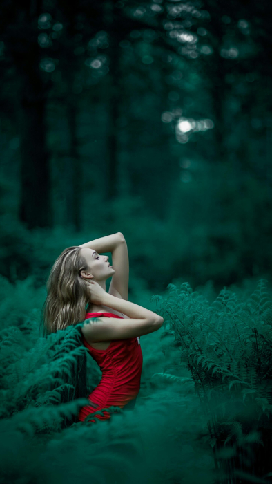 A woman poses in a vibrant red dress amidst lush green ferns in an enchanting forest setting.
