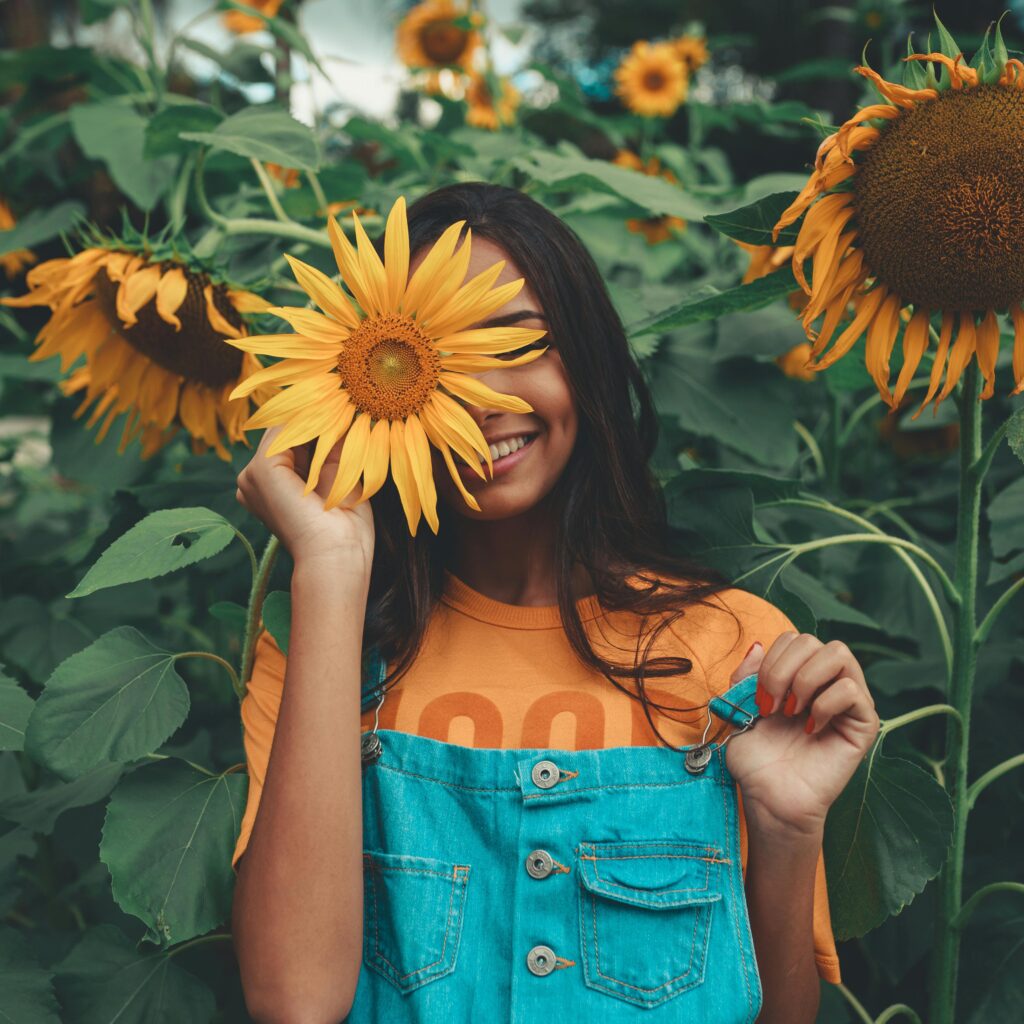 Joyful woman in denim holding sunflower among vibrant blooms in summer garden.
