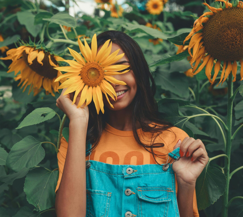 Joyful woman in denim holding sunflower among vibrant blooms in summer garden.